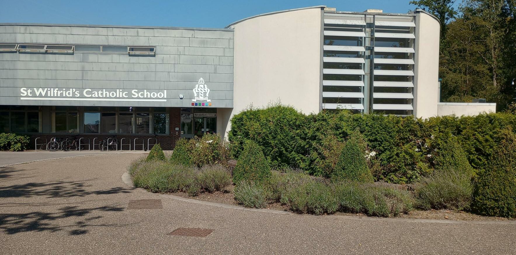 The front of St Wilfreds Catholic School, a light grey cement building with green shrubs to the right of the image