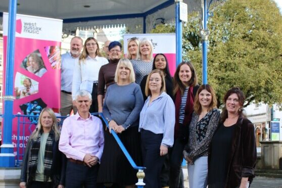 A Group Of 14 People Stand On A Bandstand, Smiling. They Have A Banner Behind Them With Their West Sussex Music Logo On.