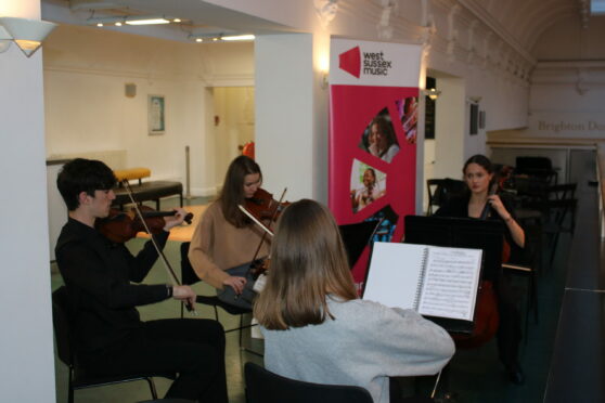 Two Young People Play Violin, Another A Cello And The Fourth Has Their Back To The Camera. A Banner In The Back Says West Sussex Music.