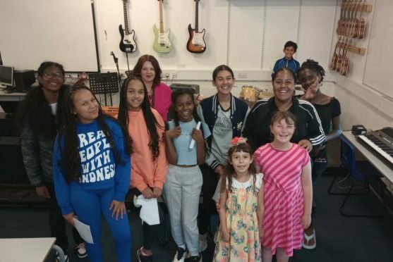 A Group Of Eleven Children And Young People Smile In A Group At The Camera. In The Background Are Guitars Hanging On The Walls, Suggesting It's A Music Room