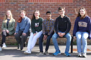 Six young people sit on a bench in front of a brick wall