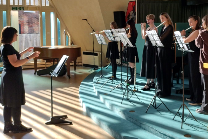 A woman in black on the left conducts five women playing flutes